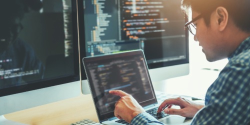 A programmer coding on a laptop and two large monitors.